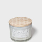 Vanilla-scented candle jar with floral lid and 'SAND+FOG' label