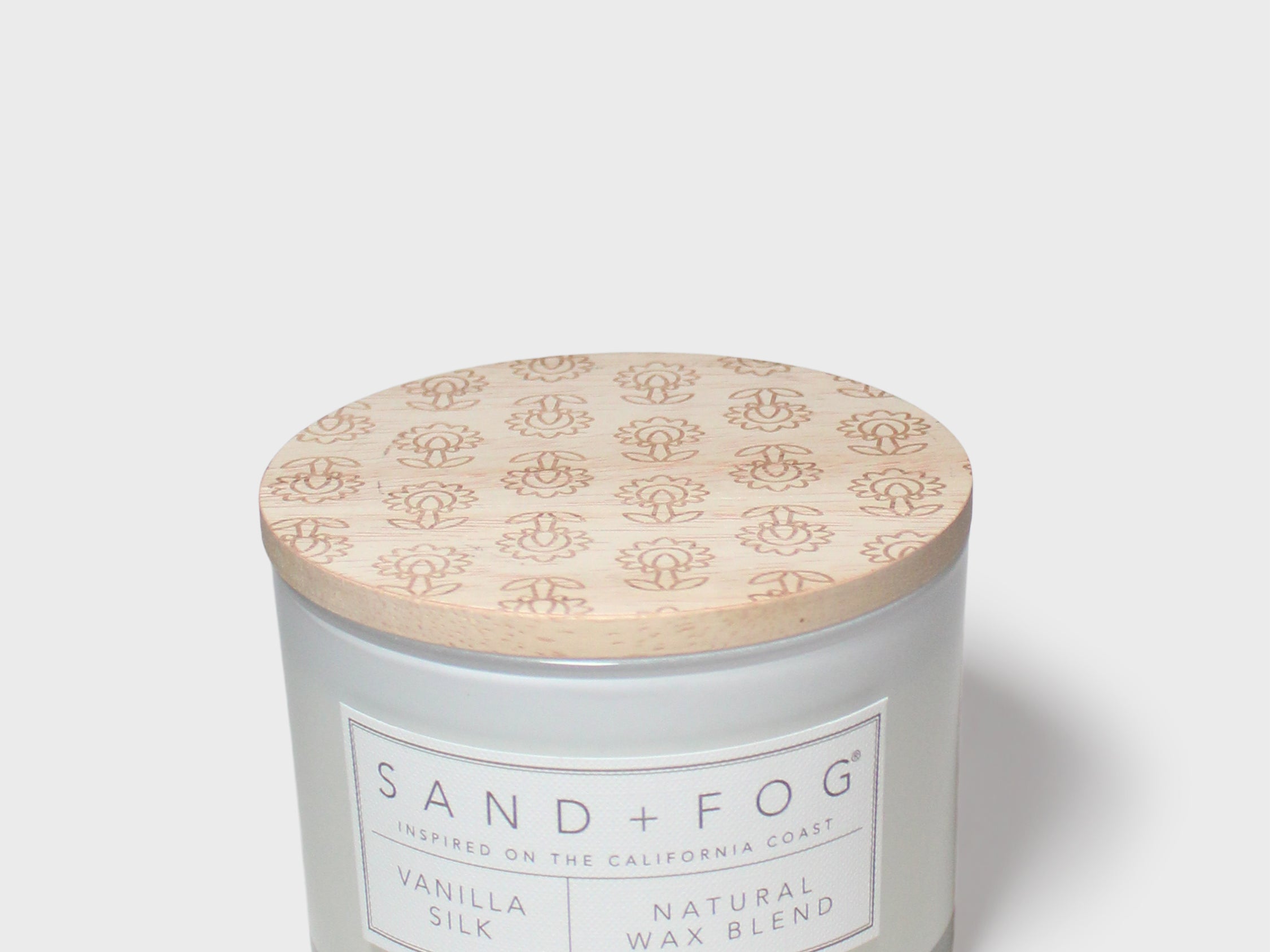 Vanilla-scented candle jar with floral lid and 'SAND+FOG' label