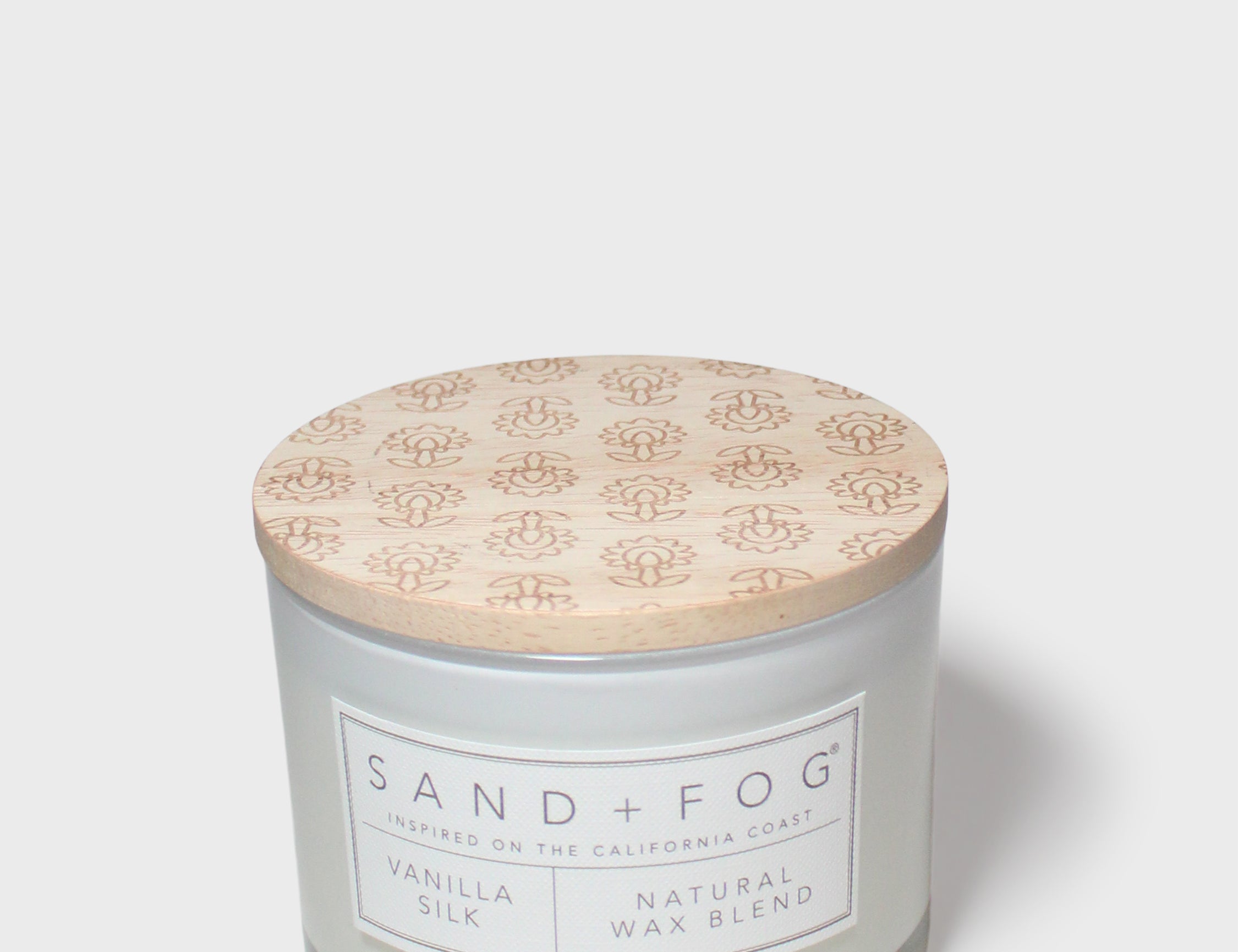 Vanilla-scented candle jar with floral lid and 'SAND+FOG' label