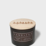 Scented candle with dark jar and light lid showing Sand+Fog Western Oak label