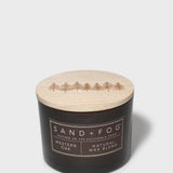 Scented candle with dark jar and light lid showing Sand+Fog Western Oak label