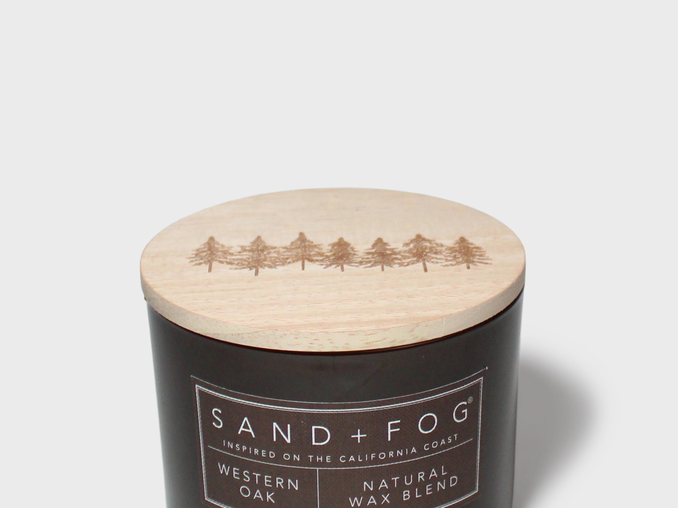 Scented candle with dark jar and light lid showing Sand+Fog Western Oak label
