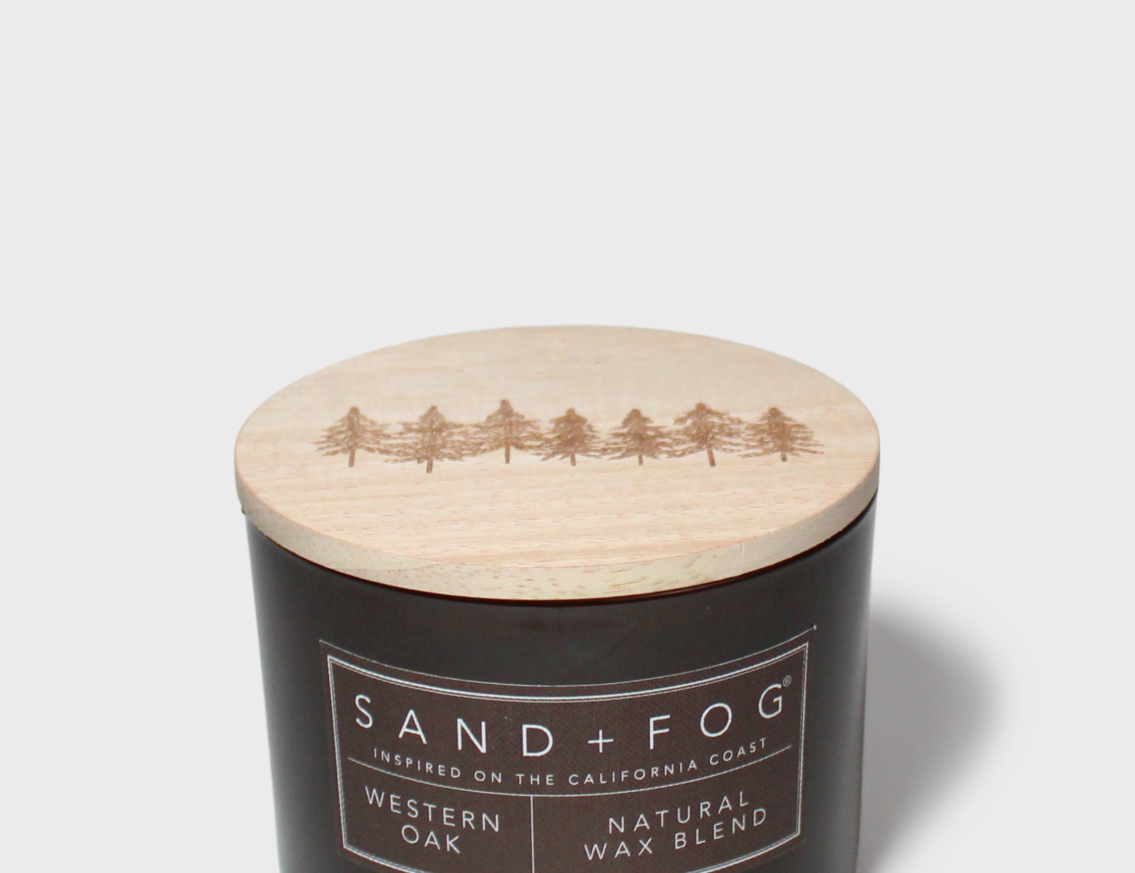 Scented candle with dark jar and light lid showing Sand+Fog Western Oak label