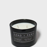 Black scented candle in glass jar, three wicks, Sand + Paws label and cream scent text