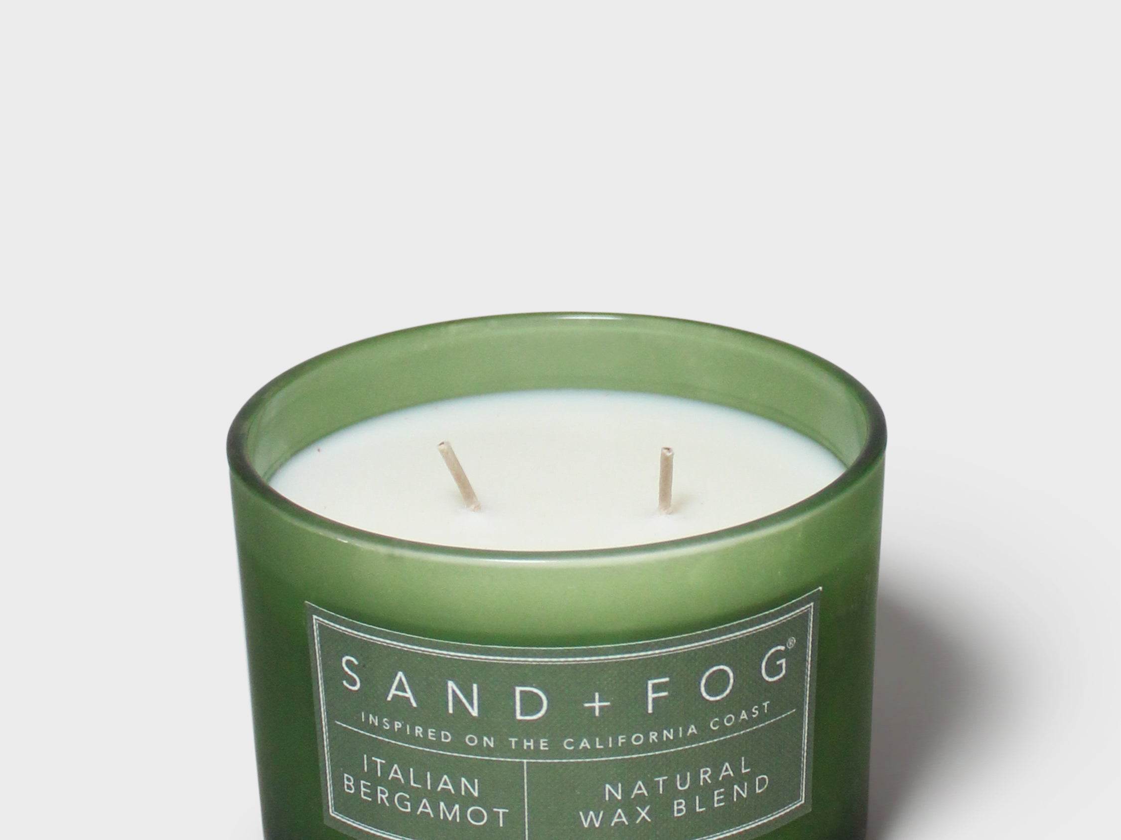 Green glass scented candle with white wax, two wicks, and SAND+FOG label