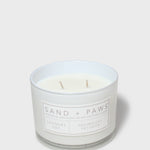 Scented candle in clear glass jar with white wax, two wicks, and Sand + Paws label