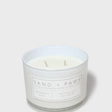 Scented candle in clear glass jar with white wax, two wicks, and Sand + Paws label