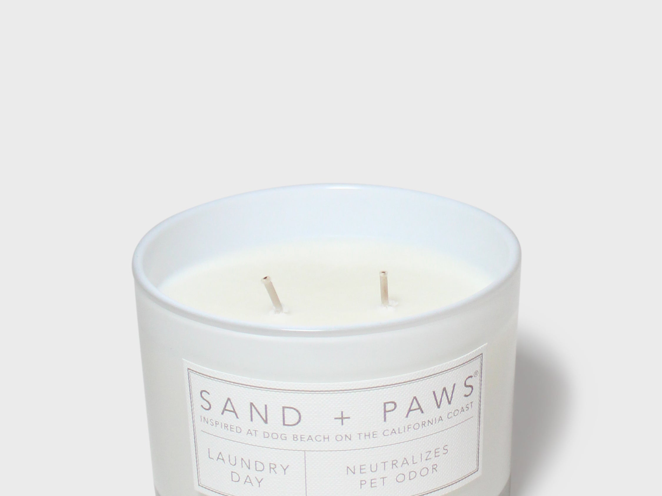 Scented candle in clear glass jar with white wax, two wicks, and Sand + Paws label