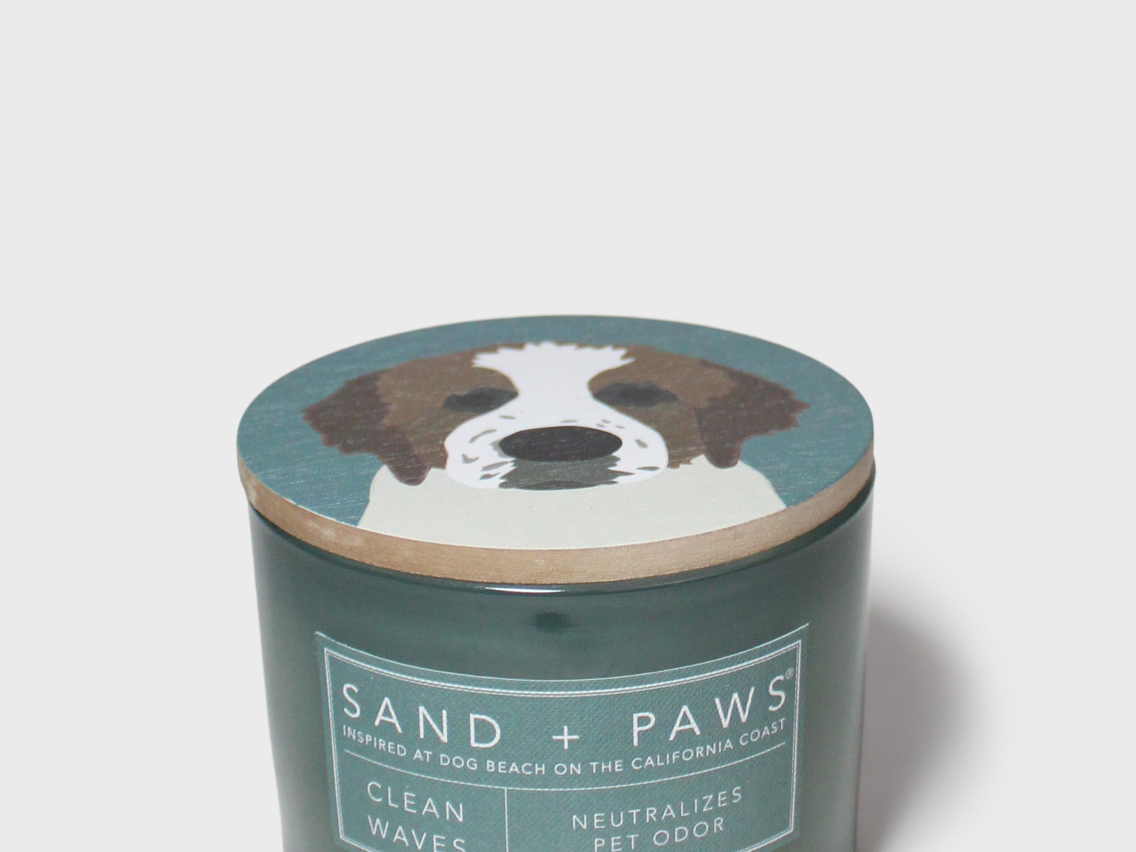 Scented candle in dark teal glass jar with "Sand + Paws" dog graphic lid