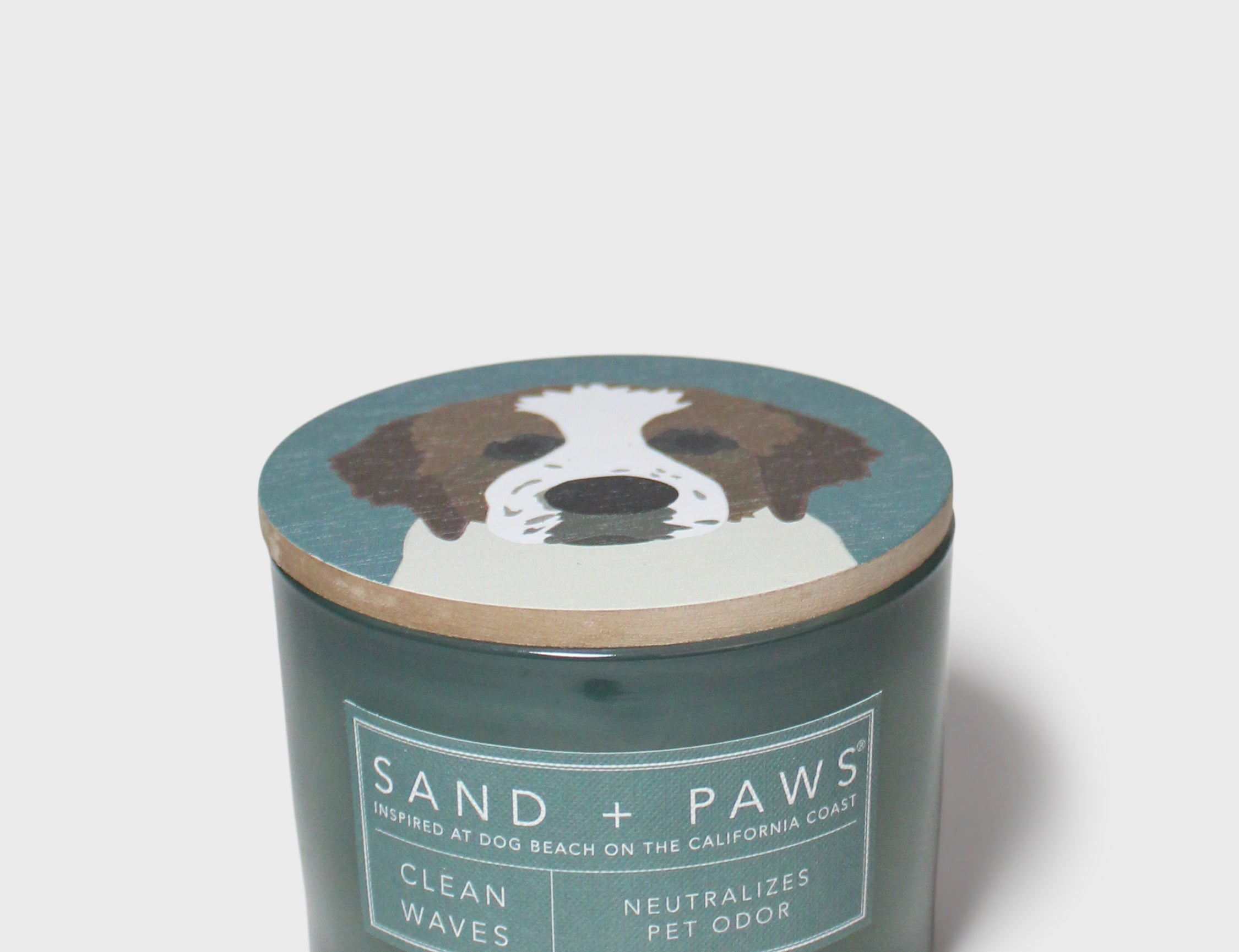 Scented candle in dark teal glass jar with "Sand + Paws" dog graphic lid