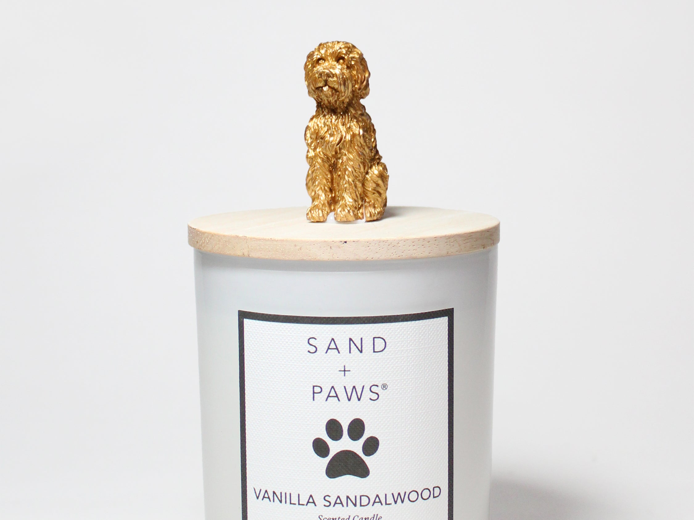 Scented candle jar with Sand + Paws Vanilla Sandalwood label and golden dog topper