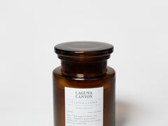 Amber glass scented candle in jar with black lid and Sand+Fog label