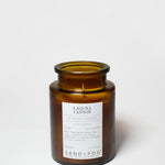 Amber glass scented candle jar with open top, visible wick, and Sand+Fog label
