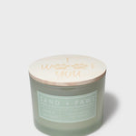 Sage green scented candle in glass jar with printed “Sand + Paws” label