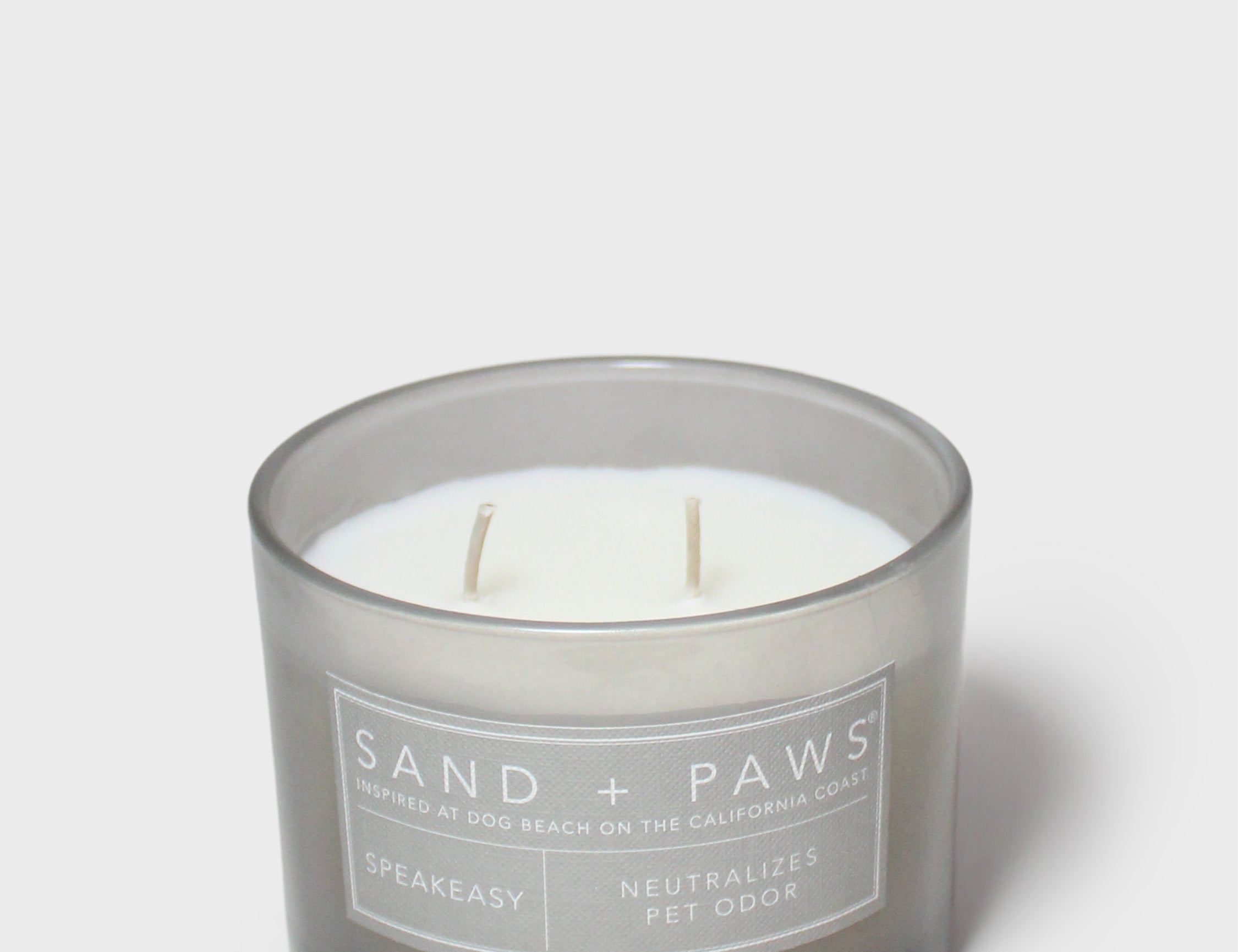 Gray glass jar scented candle with white wick and 'Sand + Paws' Speakeasy label