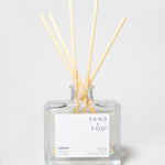 SAND + FOG Library reed diffuser with pale wooden reeds in a clear glass bottle.