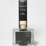 Teakwood reed diffuser in a clear glass bottle with a wood cap beside its black box.