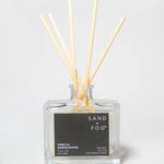 Vanilla Sandalwood reed diffuser in a clear square glass bottle with pale reeds and a black label.