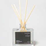 Vanilla Sandalwood reed diffuser in a clear square glass bottle with pale reeds and a black label.