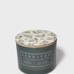 Scented candle with dark glass jar, patterned lid, and Sand + Fog Natural Wax Blend label