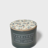 Scented candle with dark glass jar, patterned lid, and Sand + Fog Natural Wax Blend label