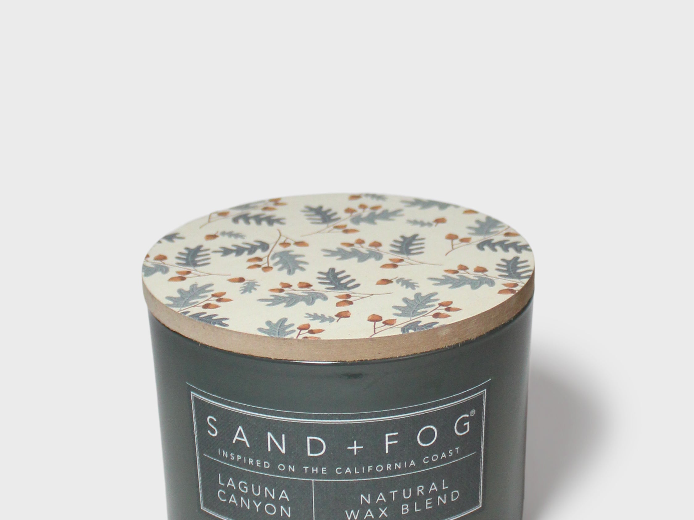 Scented candle with dark glass jar, patterned lid, and Sand + Fog Natural Wax Blend label