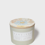 Scented candle with floral lid top and SAND+FOG label on frosted green jar