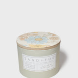 Scented candle with floral lid top and SAND+FOG label on frosted green jar