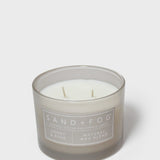Scented candle in gray glass jar with visible wicks and “SAND+FOG” label