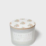 Scented candle jar with floral top and Sand + Fog wax blend label
