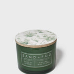 Green glass scented candle with botanical-printed lid and Sand+Fog label