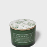 Green glass scented candle with botanical-printed lid and Sand+Fog label