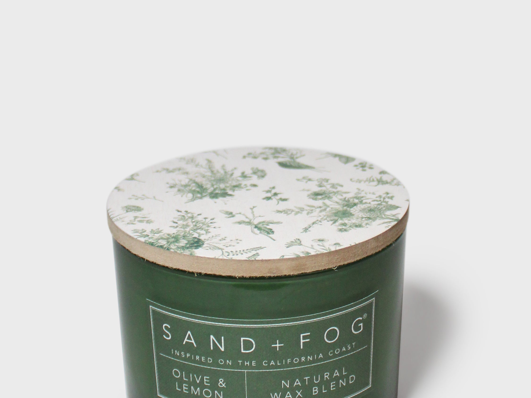 Green glass scented candle with botanical-printed lid and Sand+Fog label