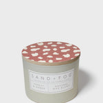 Scented candle in pale gray-green jar with red speckled lid and brand label